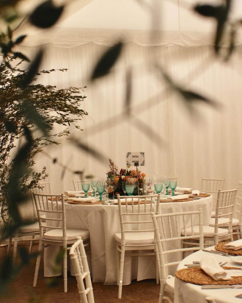 Mesa redonda decorada con cristalería verde, centro floral y sillas blancas, vista a través de ramas, en ambiente íntimo de celebración.