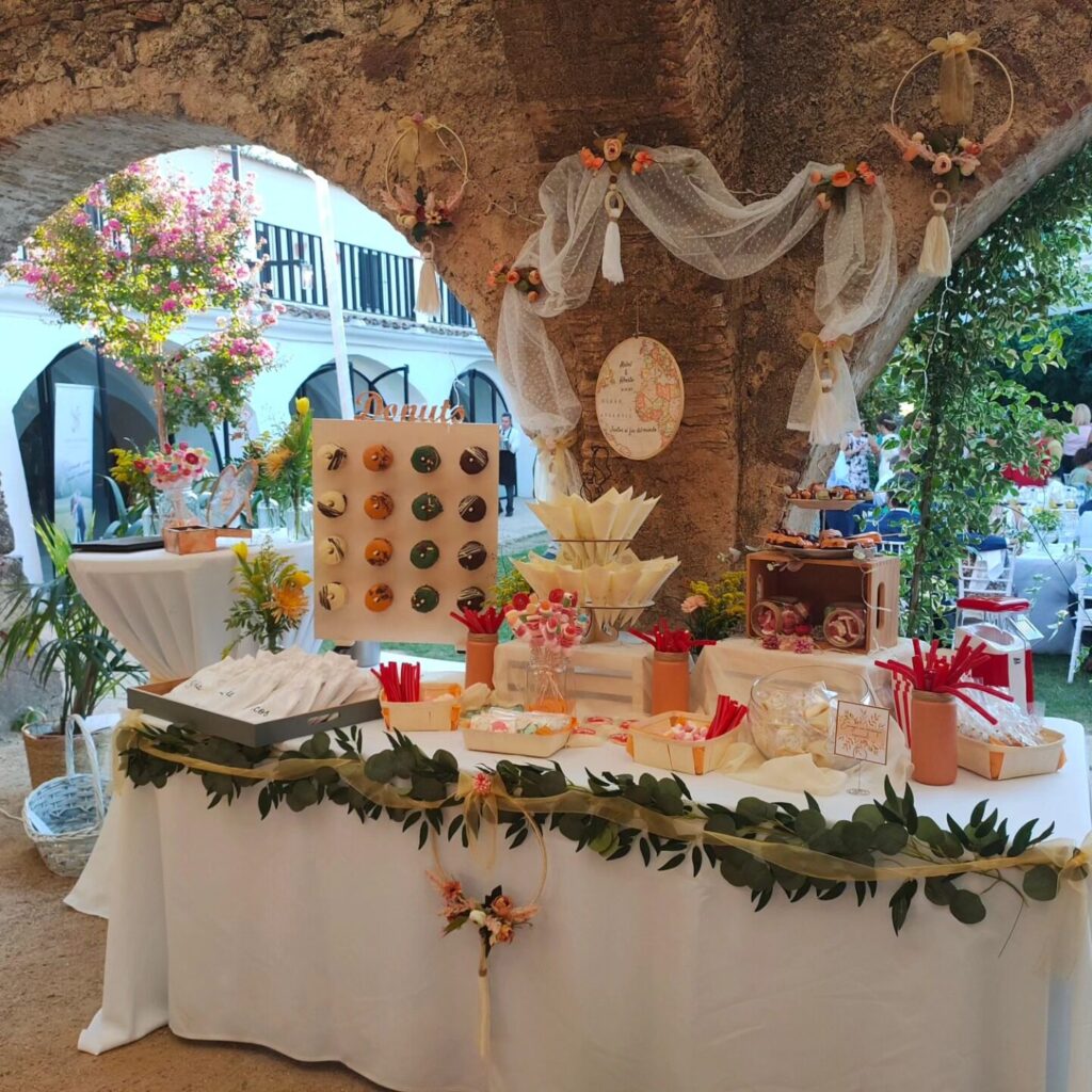 Mesa dulce decorada con detalles florales, donuts en soporte vertical, golosinas y conos de papel para candy bar de boda.